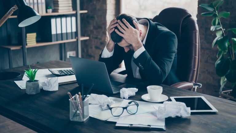 Photo of helpless business guy crumple papers holding head hands need rest work day night workaholic tired powerless wear black blazer shirt suit sitting chair office indoors
